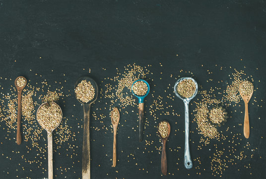 Flat-lay of various old vintage kitchen spoons full of green uncooked buckwheat grains over black stone background, top view, copy space, horizontal composition. Rustic cooking concept