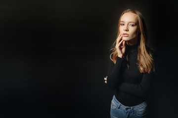 Fototapeta na wymiar Closeup portrait of smiling teen female in black pullover with hand on chin, over dark studio background. Space for text.