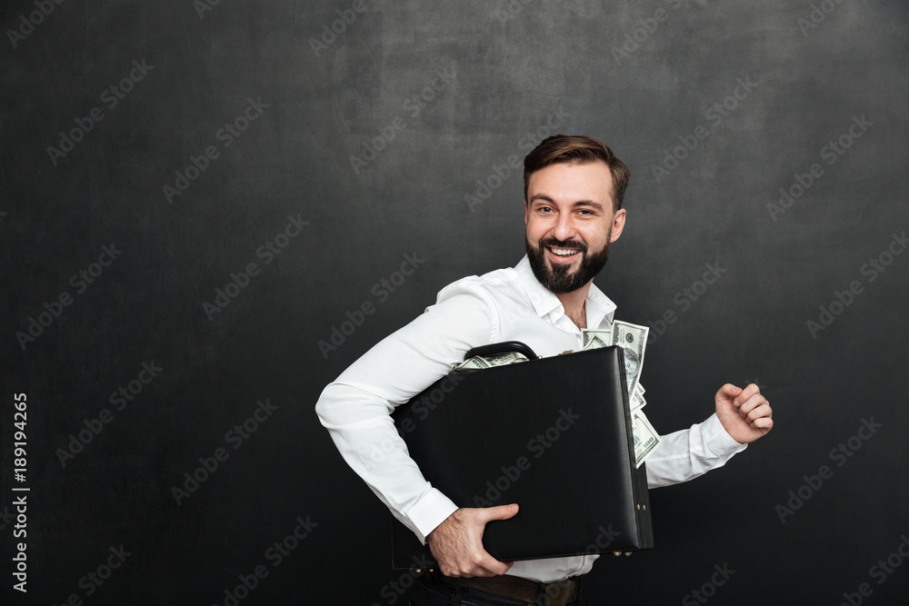 Poster funny picture of lucky man holding black briefcase full of dollar banknotes inside, isolated over da