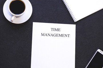 Stylish office table desk, flat lay, top view. Workplace with a white cup of coffee, a notepad, smartphone and a paper with printed Time Management inscription, black concrete background, copy space