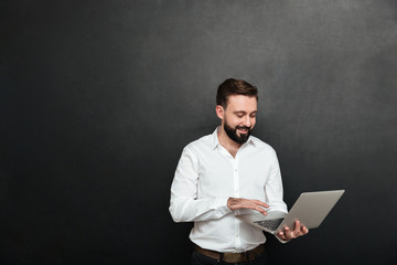 Portrait of handsome brunette man working in office using silver laptop, isolated over dark gray wall