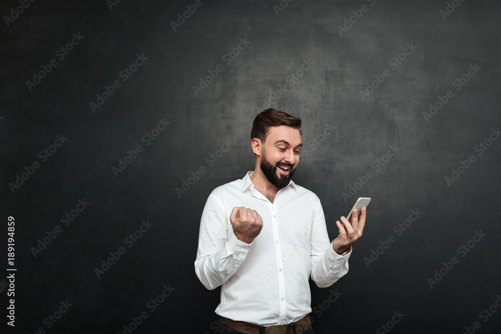 Poster delighted businessman in white shirt acting like winner clenching fist while using cellphone, over g