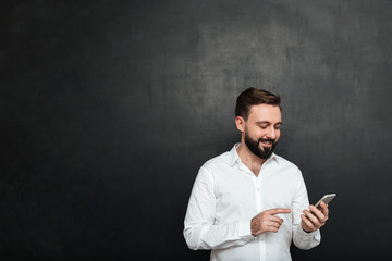 Content smiling man in white shirt typing text message or scrolling feed in social network using smartphone, over dark gray background