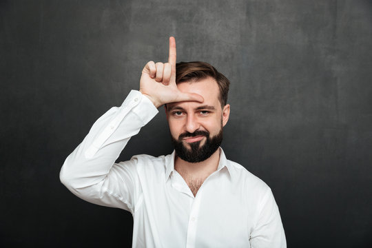 Portrait of unhappy guy 30s posing on camera and showing loser sign on his forehead, over graphite background