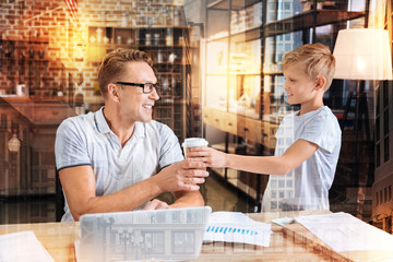 Thank you. Cheerful attentive kind boy looking at his busy hardworking father and smiling while giving him a glass of tasty coffee