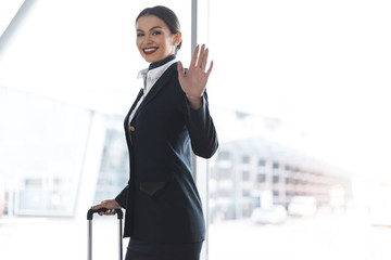 attractive young stewardess with suitcase waving at camera in airport