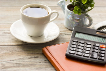 Stylish Business Office or Home Workplace Concept with a cup of black coffee, a plant in a pot and a calcilator on a brown diary on a light wooden table, close up