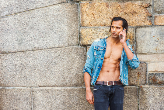 Young East Indian American Man wearing blue fashionable Denim jacket, opened, showing half naked strong body, black jeans, sneakers, standing against rock wall in New York, sad, tired, thinking..