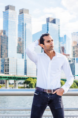 Fototapeta na wymiar Young East Indian American Man wearing white shirt, black jeans, standing in business district with high buildings by Hudson River in New York, hand on back of head, looking around, thinking..