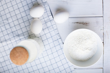 Baking Homemade Pastry Concept. A bowl of flour, eggs and a glass bottle of milk with cork. White wood and white plaid napkin as a background, top view