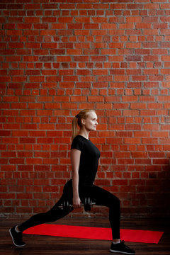 Portrait of beautiful blonde  woman in black sportswear standing against the background of a red brick wall after fitness workout.Fites concept. The girl trains with dumbbells, does attacks