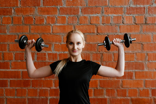 Portrait of beautiful blonde  woman in black sportswear standing against the background of  brick wall after fitness workout.Fites concept. The girl trains with dumbbells, does exercises on a biceps.
