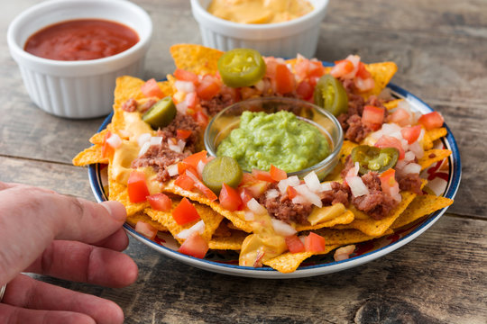 People eating mexican nachos with beef, guacamole, cheese sauce, peppers, tomato and onion in plate on wooden table