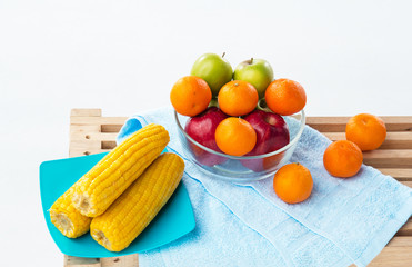 The fruit bowl with red apple,green apple and orange put beside dish of yellow corn,three oranges put beside glass bowl,on wooden timber board