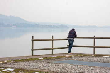 A lonely man looks at the lake leaning against a railing.