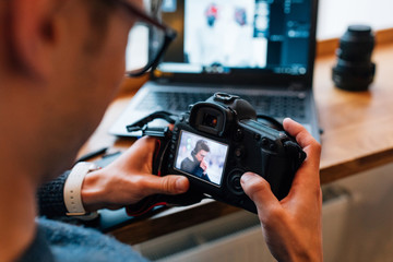 Close-up view of male hands holding professional camera, looks photos, sitting at cafe with laptop.