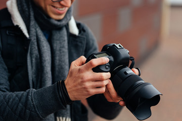 Cheerful young man looks at photos in the camera. Dressed inwarm stylish jacket, grey scarf. Standing on the street.