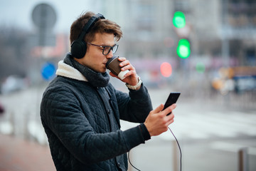 Stylish man in eyeglasses looking at mobile screen, listening to music and drinking hot coffee, standing outdoors.