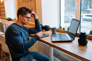 Young photographer in eyeglasses working with interactive pen display while looking at laptop screen, sitting at cafe, a cup of tea, camera and lens are on the table. Dressed in sweater and jeans.