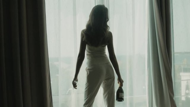 Girl with long hair in a white trouser suit holds a bottle of wine and glasses, looks out the window in a luxurious hotel room