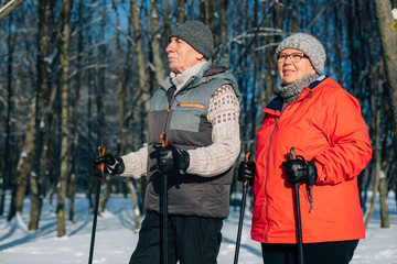 Pretty senior couple standing with nordic walking poles in winter park. Mature woman and old man...