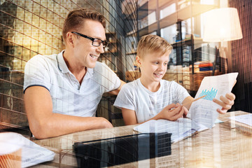 Lets compare. Cheerful kid expressing positivity while demonstrating his project and sitting near his father