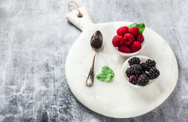 fresh summer berries raspberries and blackberries with mint on a marble serving board in small marble bowls on a stone table. dessert serving of healthy food