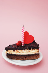 Piece of Chocolate Cake with Candle and Decorative Hearts on top. Pink Background with copy space. Vertical image.