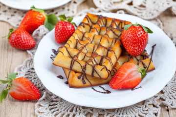 Belgium waffles with strawberries and chocolate decoration on plate with cup of coffee and small jar with chocolate on wooden table, selective focus