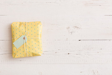 Top view on small wrapped yellow gift with ribbons and tag on white wooden background. Present for birthday, Christmas or any other celebration or party