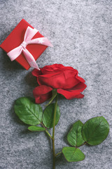One red rose and a cute gift box - Gifting theme image with a beautiful red rose and a cute little gift box wrapped in red paper and a pink ribbon with tied bow, on a grey background.
