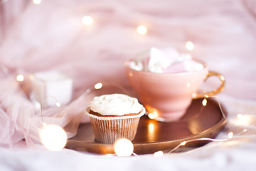 Cupcake with creamy top, cup of coffee on tray in bed over lights at background. Selective focus. Good morning.