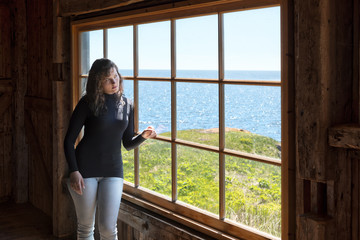 Side of one, lonely, alone young woman standing by large glass window looking at peaceful ocean view, cliff, in rustic wooden house