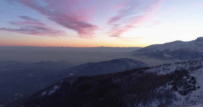 Backward aerial top view over winter snowy mountain and woods forest at sunset or sunrise.Blue hour dusk or dawn twilight Alps mountains snow season establisher.4k drone flight establishing shot
