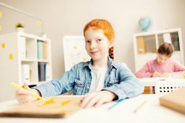 Smiling schoolgirl in denim jacket looking at camera at lesson of drawing