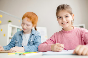 Cute girl with toothy smile looking at camera at lesson with her classmate near by