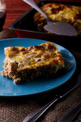 On a blue plate lies a piece of mouth-watering meat casserole. A dish with a casserole and a spatula for eating in the background. Photo in the kitchen while cooking food.