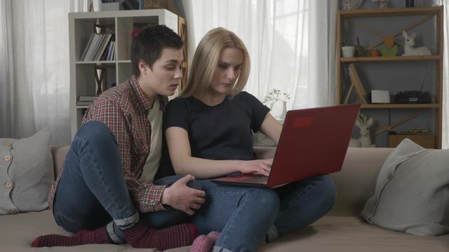 Two young lesbian girls are sitting on the couch, using a computer, scrolling the internet, shopping online, the blonde maliciously closes the laptop, quarrel 60 fps