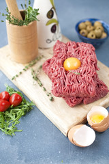 Fresh beef meat in forcemeat, on a wooden board. Raw eggs and tomatoes are small. Herbs for cooking. On a gray stone background. Free space for writing text.