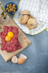 Fresh beef meat in forcemeat, on a wooden board. Raw eggs and tomatoes are small. Herbs for cooking and round buns. On a gray stone background. Free space for writing text.