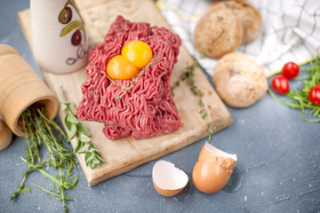 Fresh beef meat in forcemeat, on a wooden board. Raw eggs and tomatoes are small. Herbs for cooking and round buns. On a gray stone background. Free space for writing text.