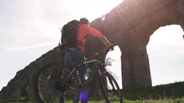 Cyclist with bike arrives under majestic ancient roman aqueduct at sunset. Young attractive athletic man with orange sportswear and backpack in parco degli acquedotti in Rome