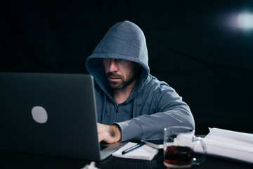 a man hacker in a sweatshirt with a hood sits behind a laptop and drinks tea