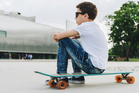 Back view. The boy, dressed in a white T-shirt and blue jeans, sits on a city street on a skateboard. On background modern building in soft focus. Lifestyle. Space for text, logo, image. Mock up.