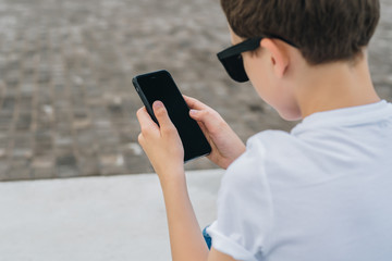 Back view. Close-up of smartphone screen in hands of boy in white T-shirt. Teenager sits outdoor and uses digital gadget, browsing internet, checking email.Computer games,social networks.Bokeh effect.