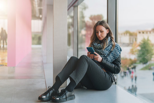 Young woman sits on floor near large window and uses smartphone.Hipster girl looking on screen of phone,blogging,chatting,browsing internet. Online marketing, education, distance work. Social network.