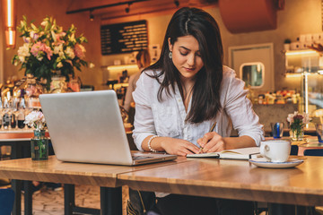 Young businesswoman is sitting in cafe at table in front of laptop, working, making notes in her notebook. Student girl learning online, writing conspectus. Online marketing, education, e-learning.