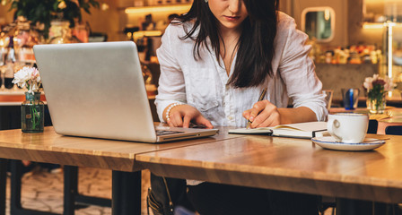 Young businesswoman is sitting in cafe at table in front of laptop, working, making notes in her notebook. Student girl learning online, writing conspectus. Online marketing, education, e-learning.
