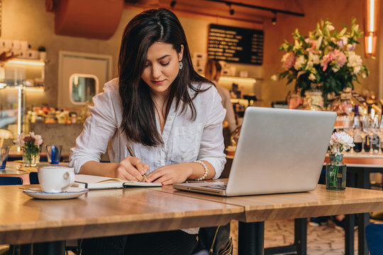 Young businesswoman is sitting in cafe at table in front of laptop, working, making notes in her notebook. Student girl learning online, writing conspectus. Online marketing, education, e-learning.