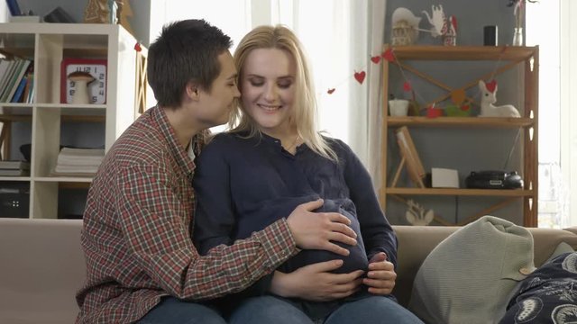 Two young lesbian girls sitting on the couch, pregnant blonde caresses the belly, waiting for the baby's appearance, coziness, love, happiness, kissing.60 fps